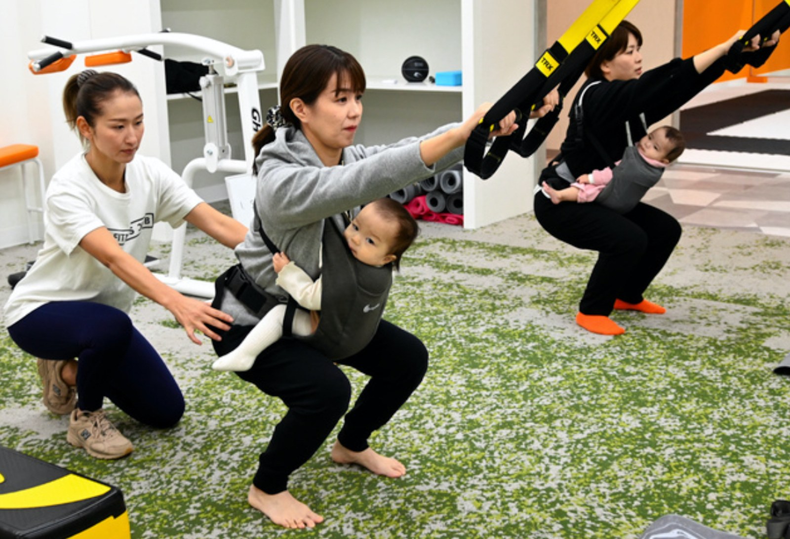 赤ちゃんを抱っこひもで抱えたままTRXでスクワットをするママとトレーナー
