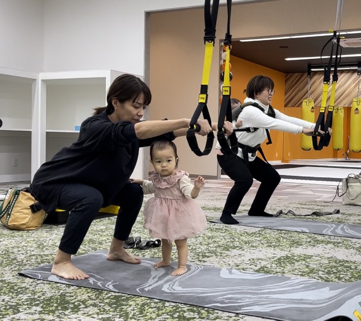 TRXでスクワットをするママの前に立つ赤ちゃん